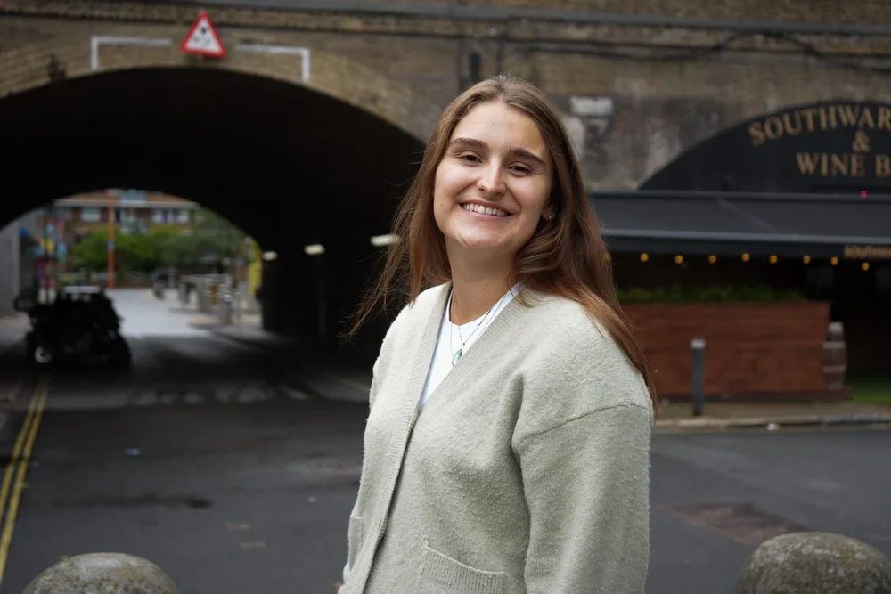 A smiling young woman with brown hair standing outdoors under a bridge, wearing a beige jacket.