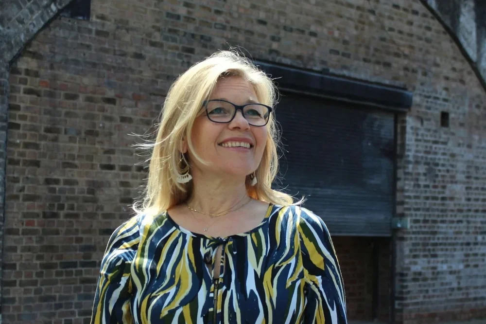 A woman with blonde hair and glasses smiling outdoors in front of a brick building.