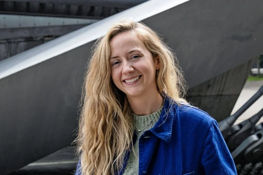 Young woman with long curly blonde hair smiling outdoors, wearing a blue jacket over a sweater.