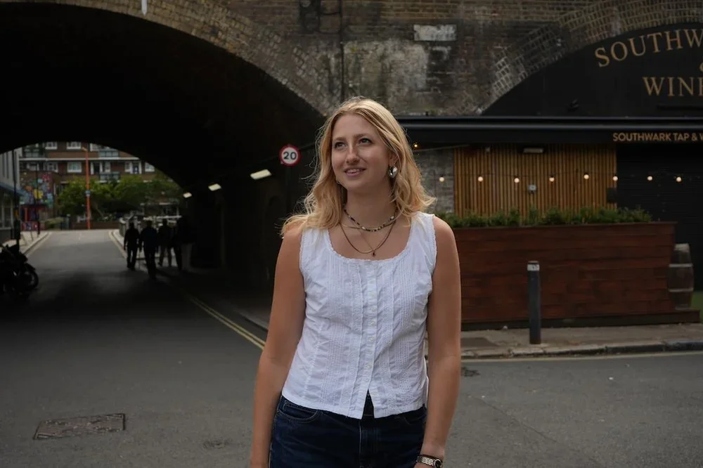 A young woman with blonde hair wearing a white sleeveless top and necklaces, standing under a bridge near a restaurant and a group of people in the background.