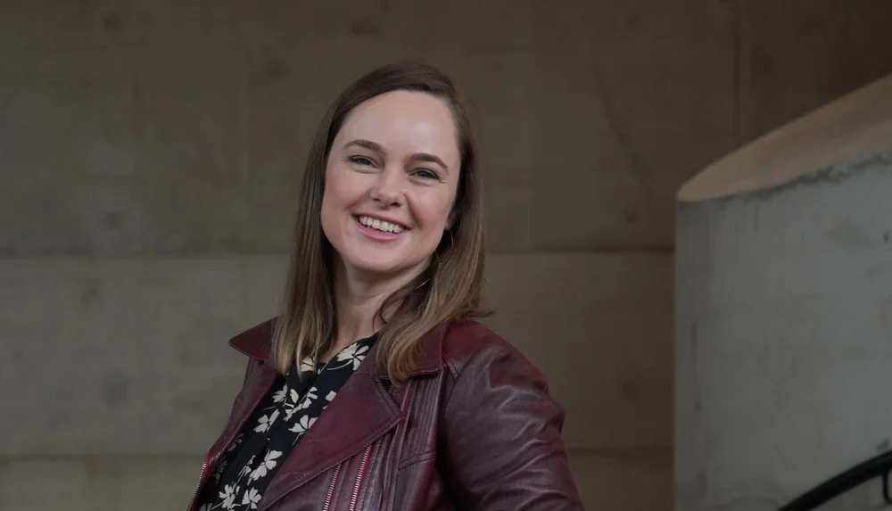 A smiling woman with shoulder-length brown hair, wearing a maroon leather jacket over a black and white floral top, standing indoors against a plain brown wall.