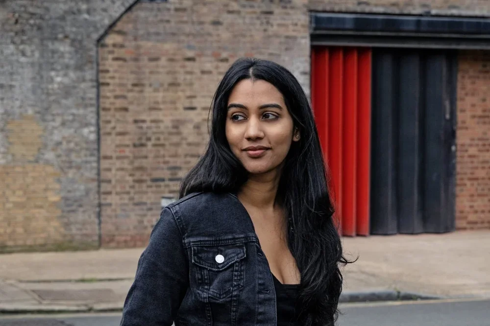 A woman with long black hair wearing a black jacket, standing outdoors in front of a brick wall with red and black doors.
