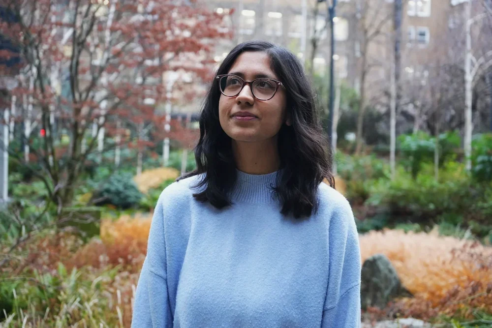 A woman with dark hair and glasses standing outdoors in a park during autumn.
