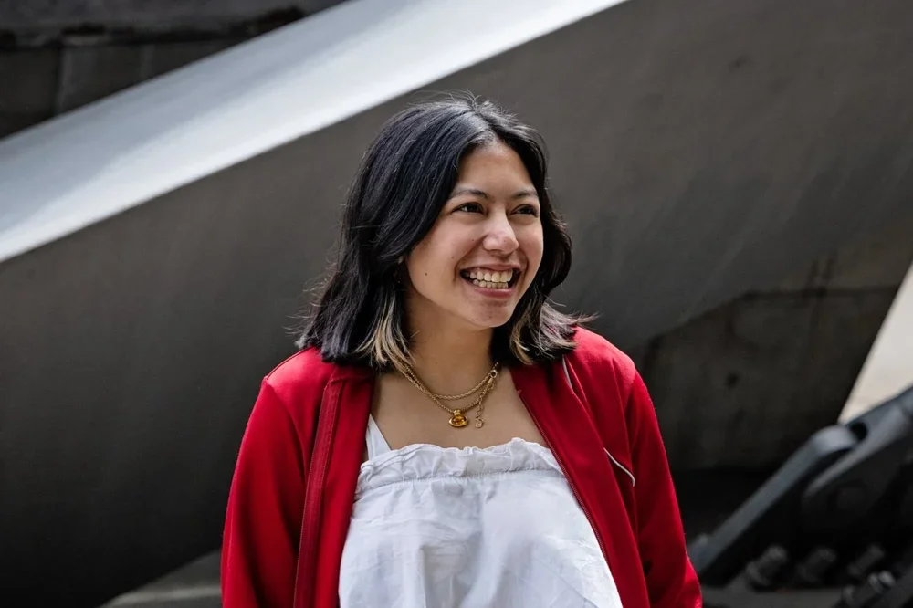 A young woman smiling outdoors, wearing a red jacket over a white top, with shoulder-length dark hair and layered necklaces.
