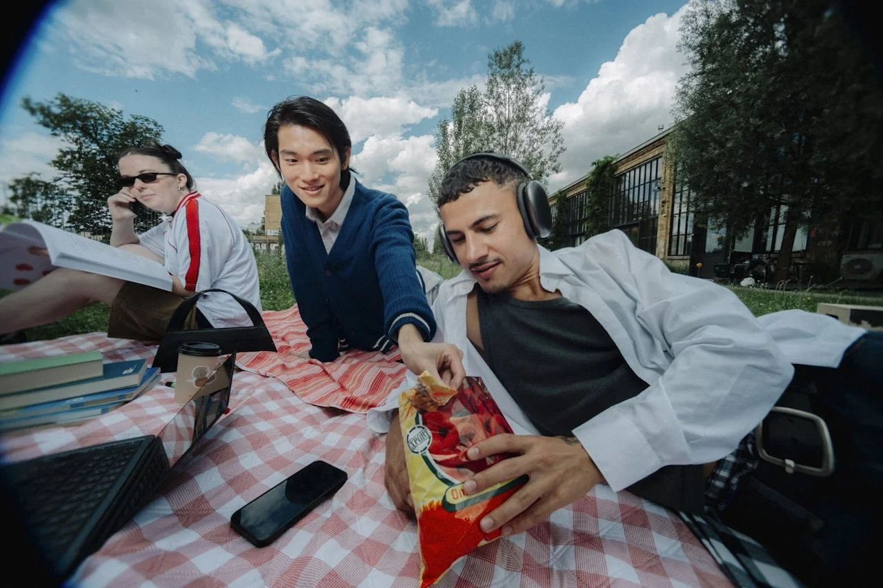 Three young people sitting on a picnic blanket outdoors, with snacks and books, enjoying a sunny day.