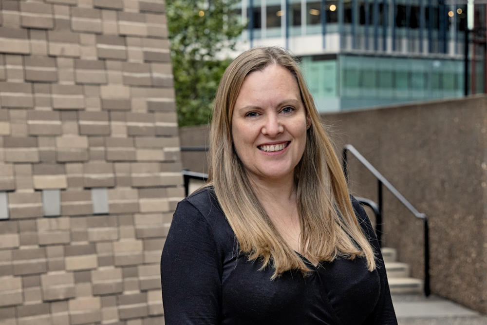 A smiling woman with long blonde hair wearing a black top outdoors near a brick building and a staircase.