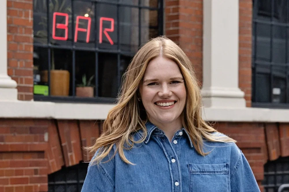 A young woman with blonde hair smiling in front of a brick building with a window that has a neon red sign spelling 'BAR'.