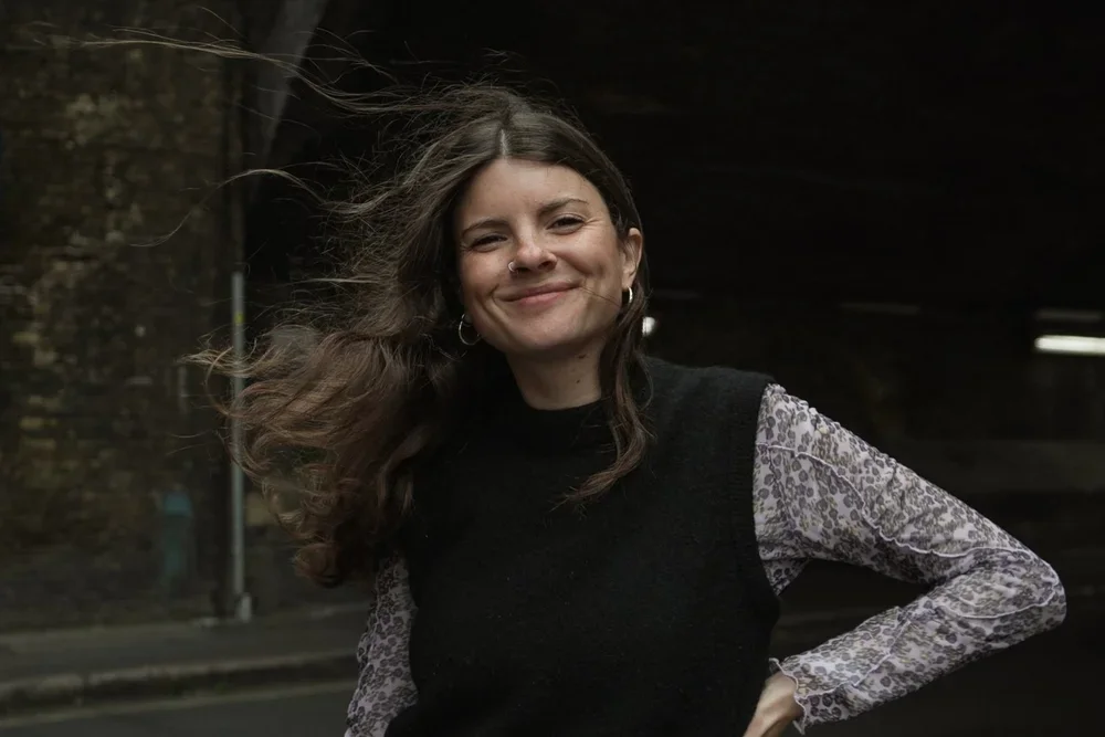 A woman with long, wavy hair smiling outdoors at night, wearing a black sleeveless vest over a leopard print long-sleeve shirt, standing with one hand on her hip.