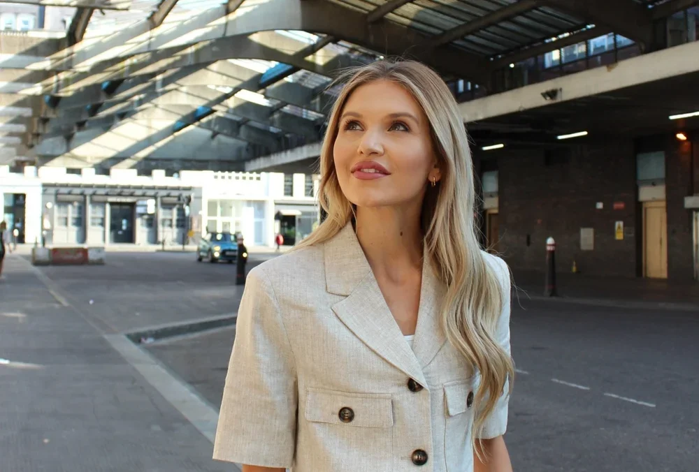 A young woman with long blonde hair, wearing a beige blazer, standing outdoors underneath a modern architectural structure with glass and steel design.