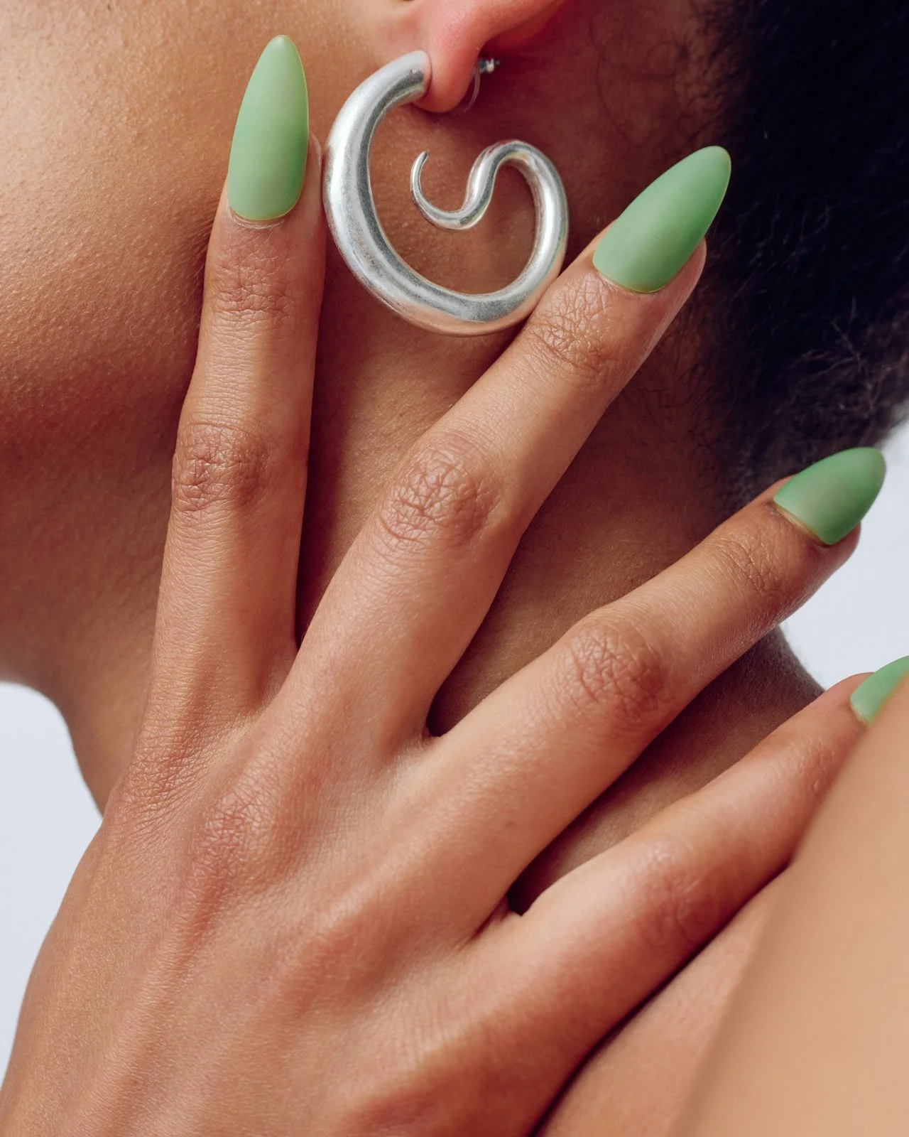 Close-up of a person with matte green nails holding a large, silver, spiral-shaped earring near their ear and cheek.