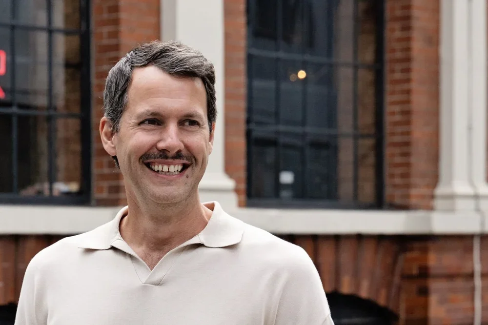 A man with dark hair and a mustache smiling outside a brick building with large windows.