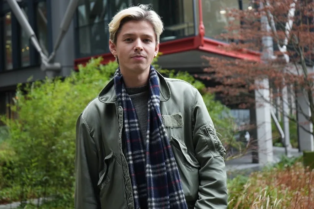 Young man with blond hair standing outdoors in front of greenery and modern buildings.