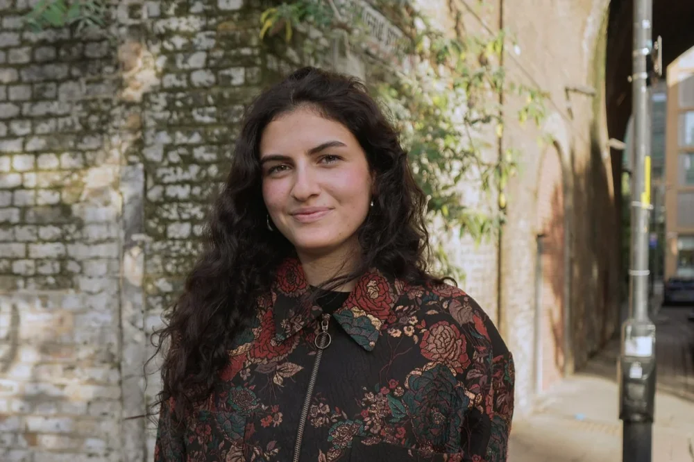 A young woman with long, curly dark hair standing outdoors in front of a rustic brick wall and some greenery, smiling at the camera.