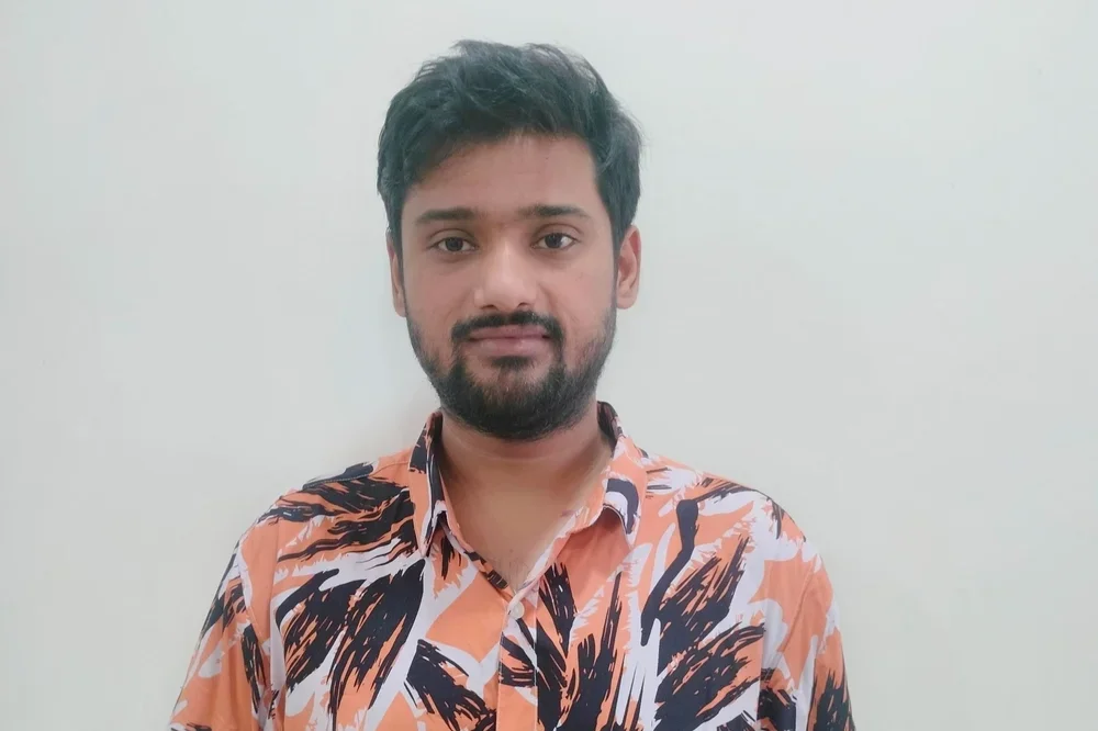 A man with a beard and short, dark hair wearing an orange and black patterned shirt standing against a plain white wall.
