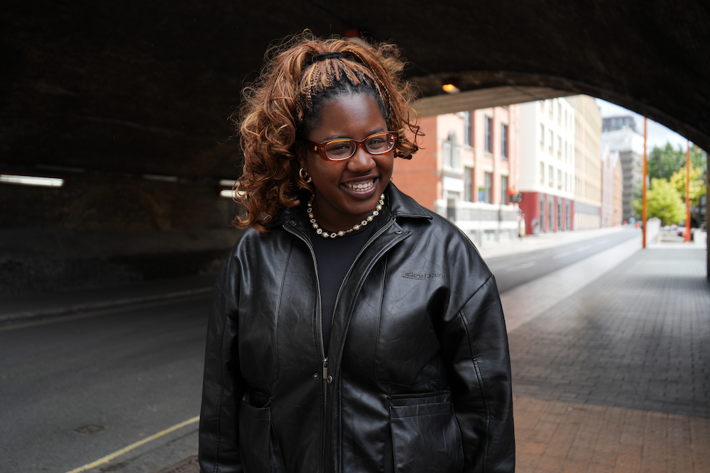 A woman with curly hair, wearing glasses, a black leather jacket, and a beaded necklace, smiles while standing under an urban bridge.