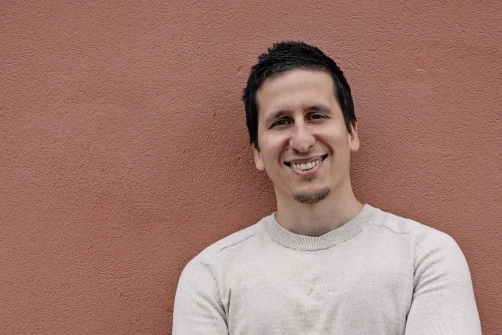 A smiling man with dark hair and a short beard, wearing a light gray shirt, standing against a reddish-brown wall.