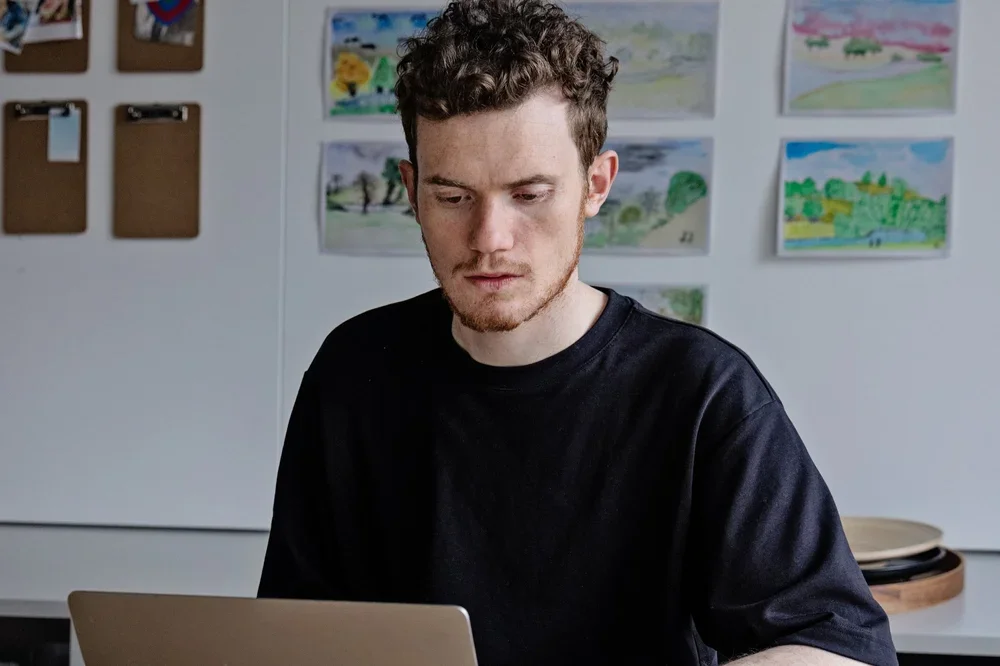 Young man with curly hair and a beard working on a laptop indoors, with children's drawings and clipboards on the wall behind him.