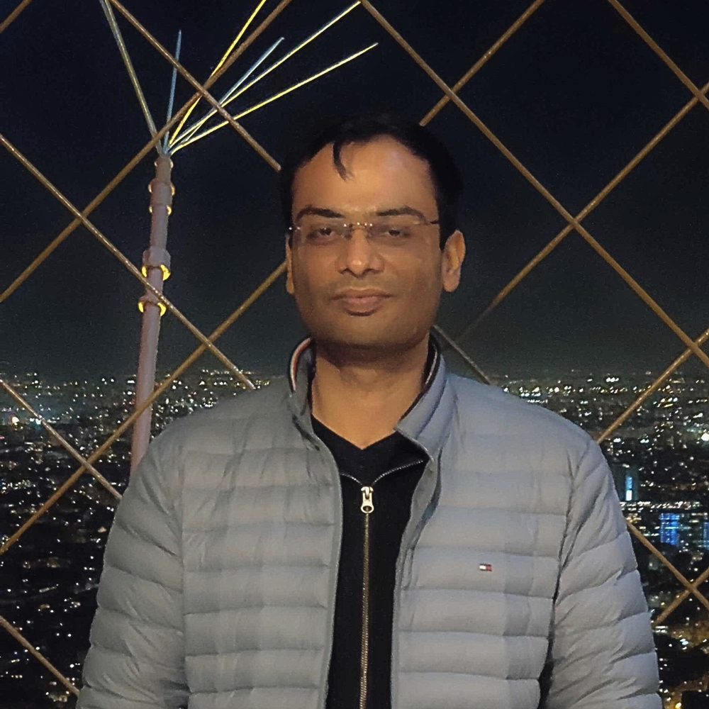 A man wearing glasses and a gray jacket standing in front of cityscape at night with lights and a metal grid structure.