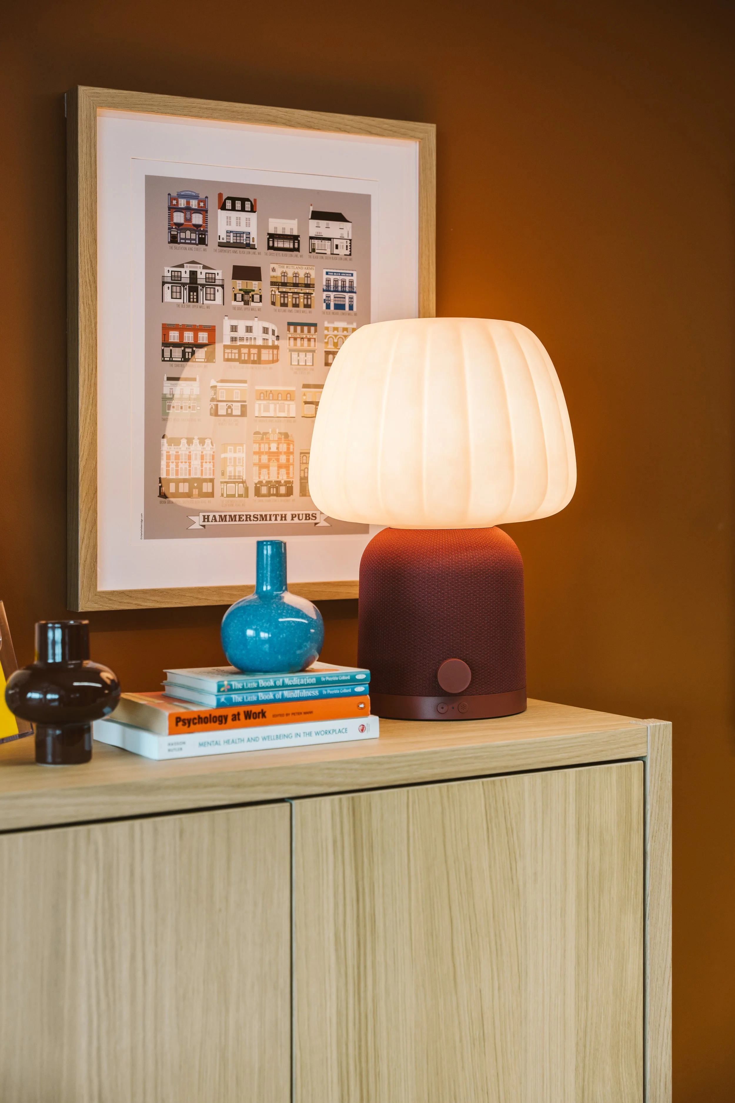 A wooden sideboard with a modern lamp, books on mental health and well-being, decorative glass bottles, and a colorful picture of various building facades hanging on a brown wall.