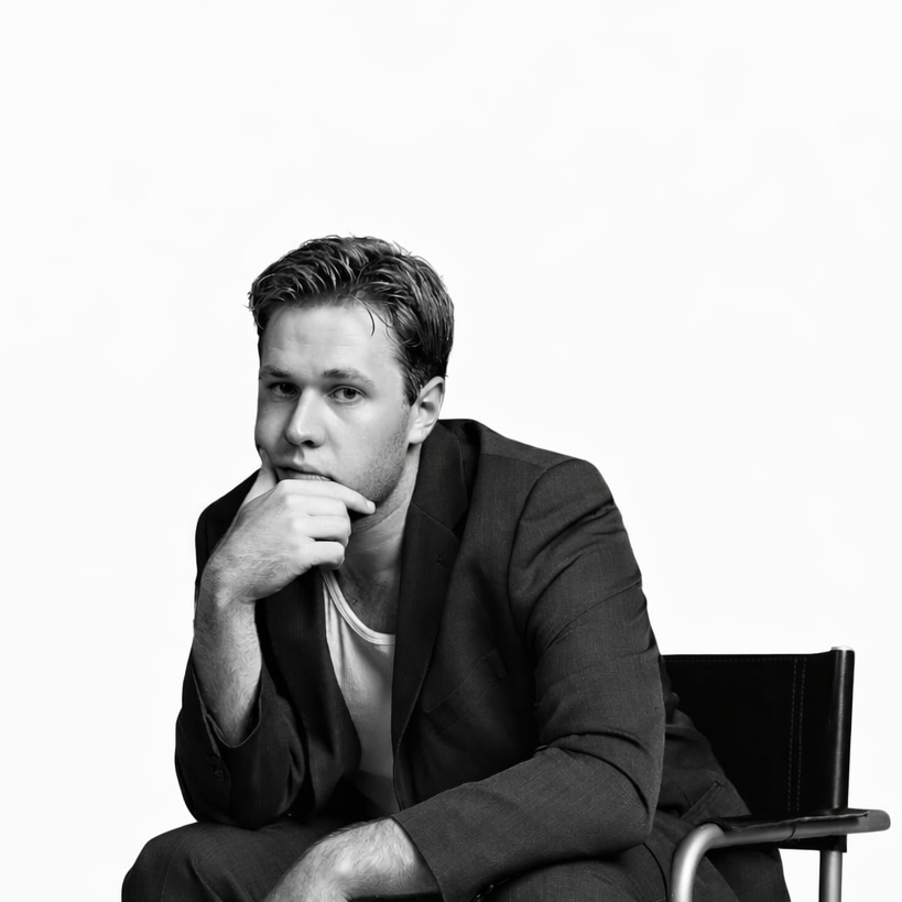 Black and white portrait of a young man sitting in a wheelchair, with his chin resting on his hand, wearing a suit jacket and a T-shirt, looking at the camera thoughtfully.
