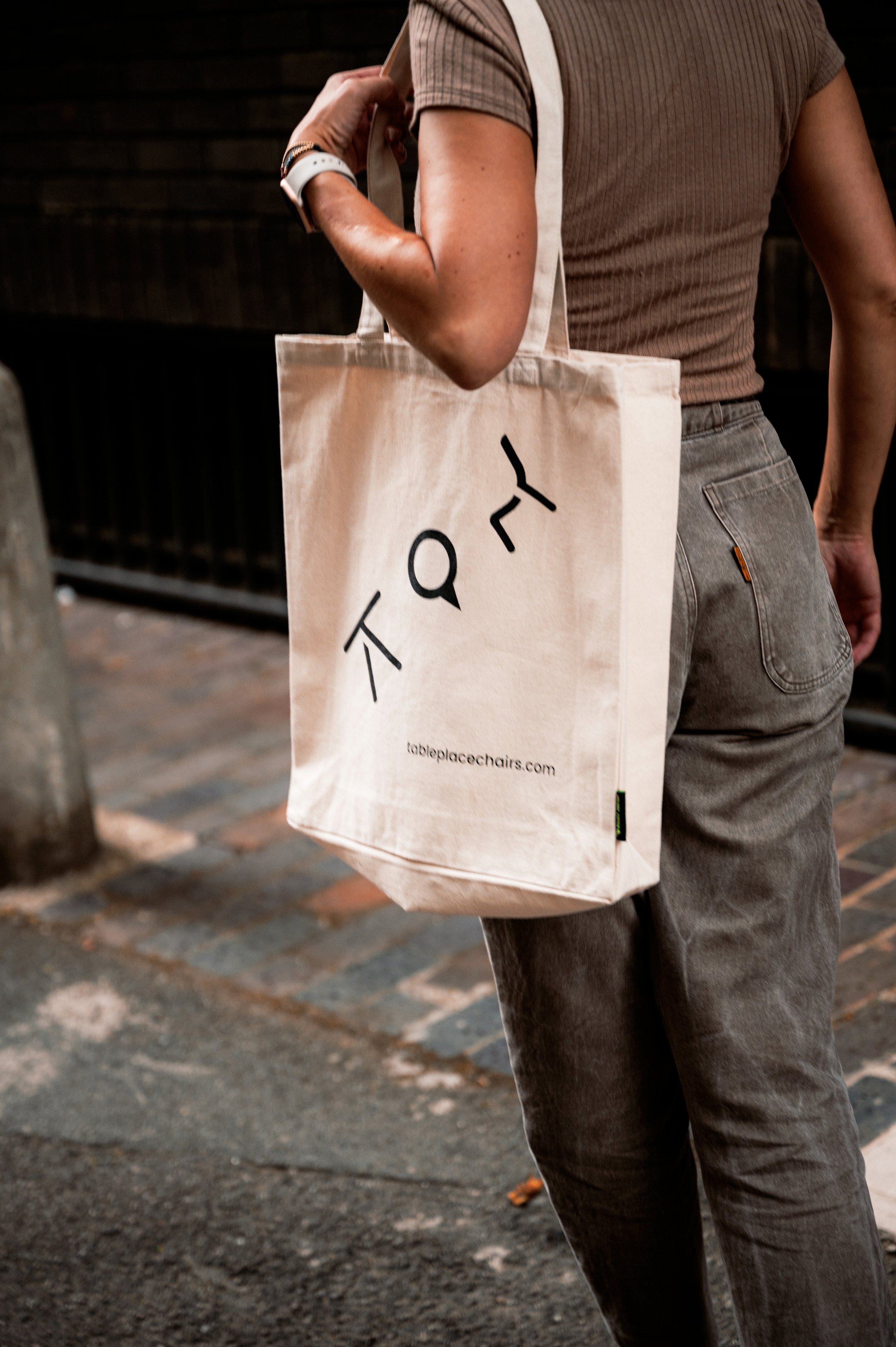 Person carrying a beige tote bag with black text on a city sidewalk.