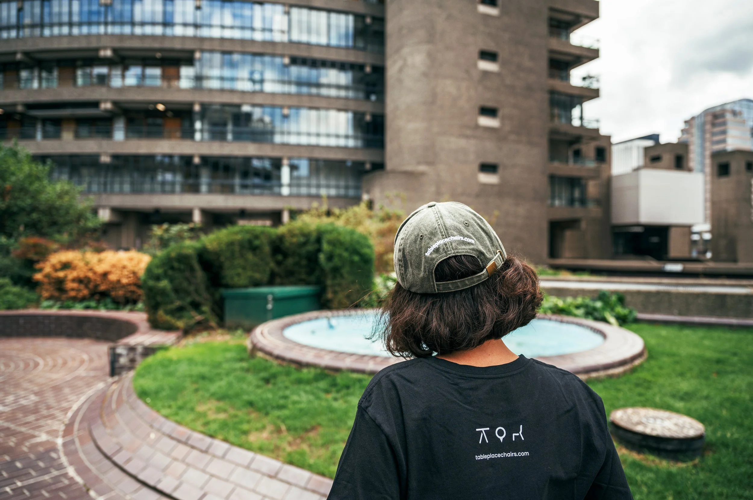 A person with shoulder-length curly hair wearing a khaki baseball cap and a black T-shirt stands outdoors on a paved brick area near a small round fountain, surrounded by green bushes and trees, with modern high-rise buildings in the background.