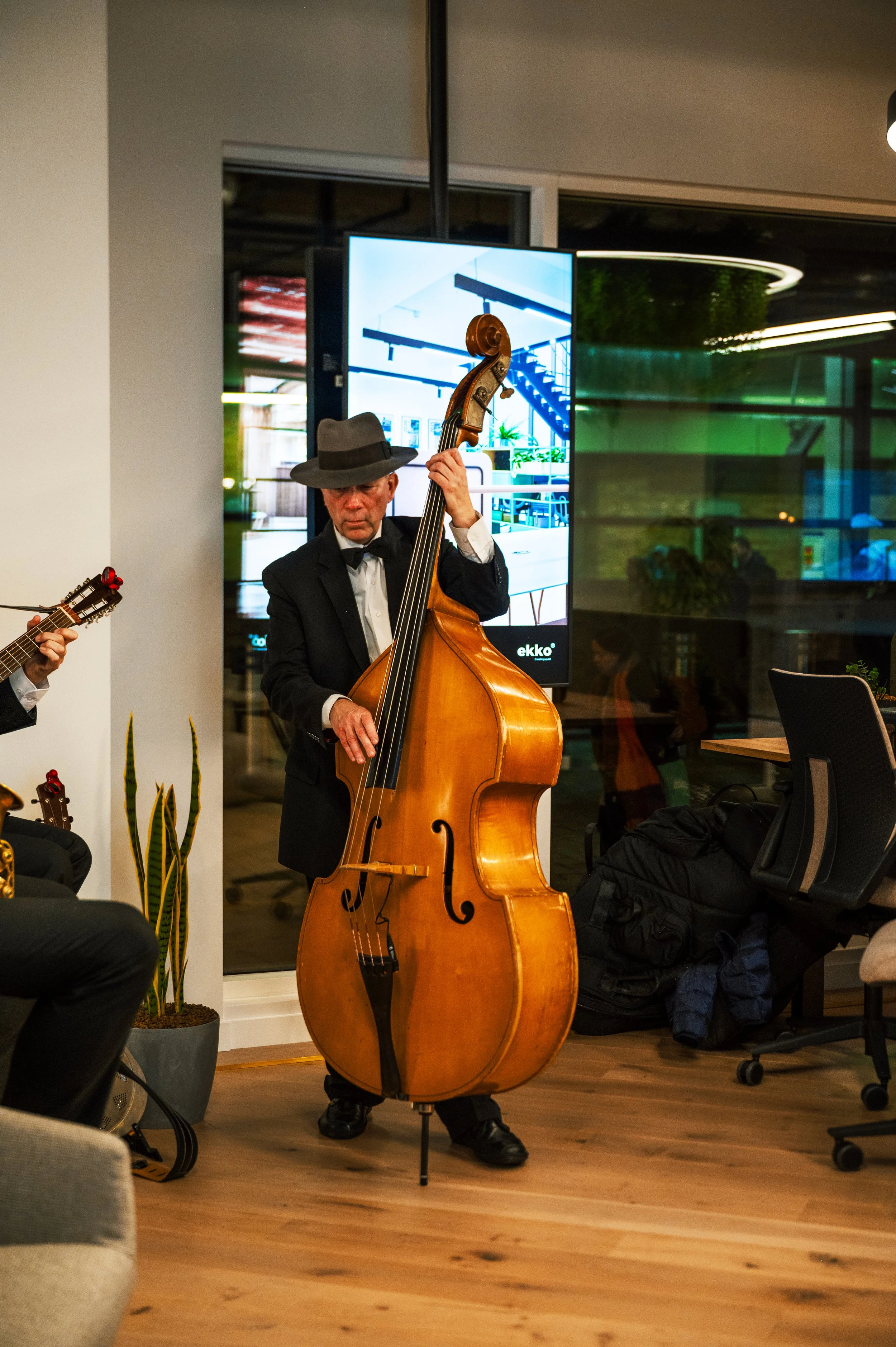 A man in a black suit, white shirt, black bow tie, and a grey fedora playing a double bass during a live music performance indoors.