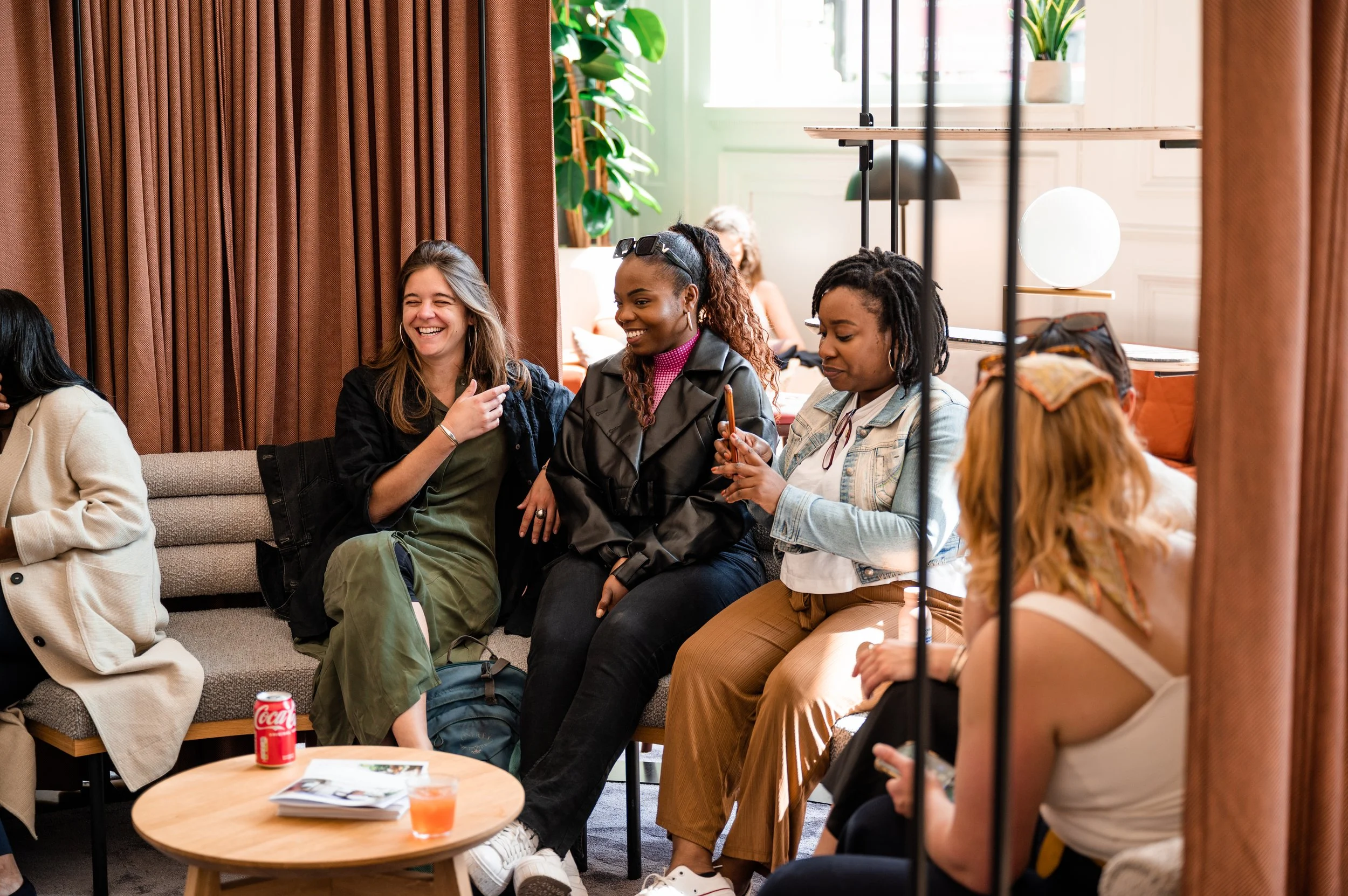 Group of women sitting on a couch and chairs, smiling, chatting, and using a phone in a modern, well-lit lounge with green plants and a window.