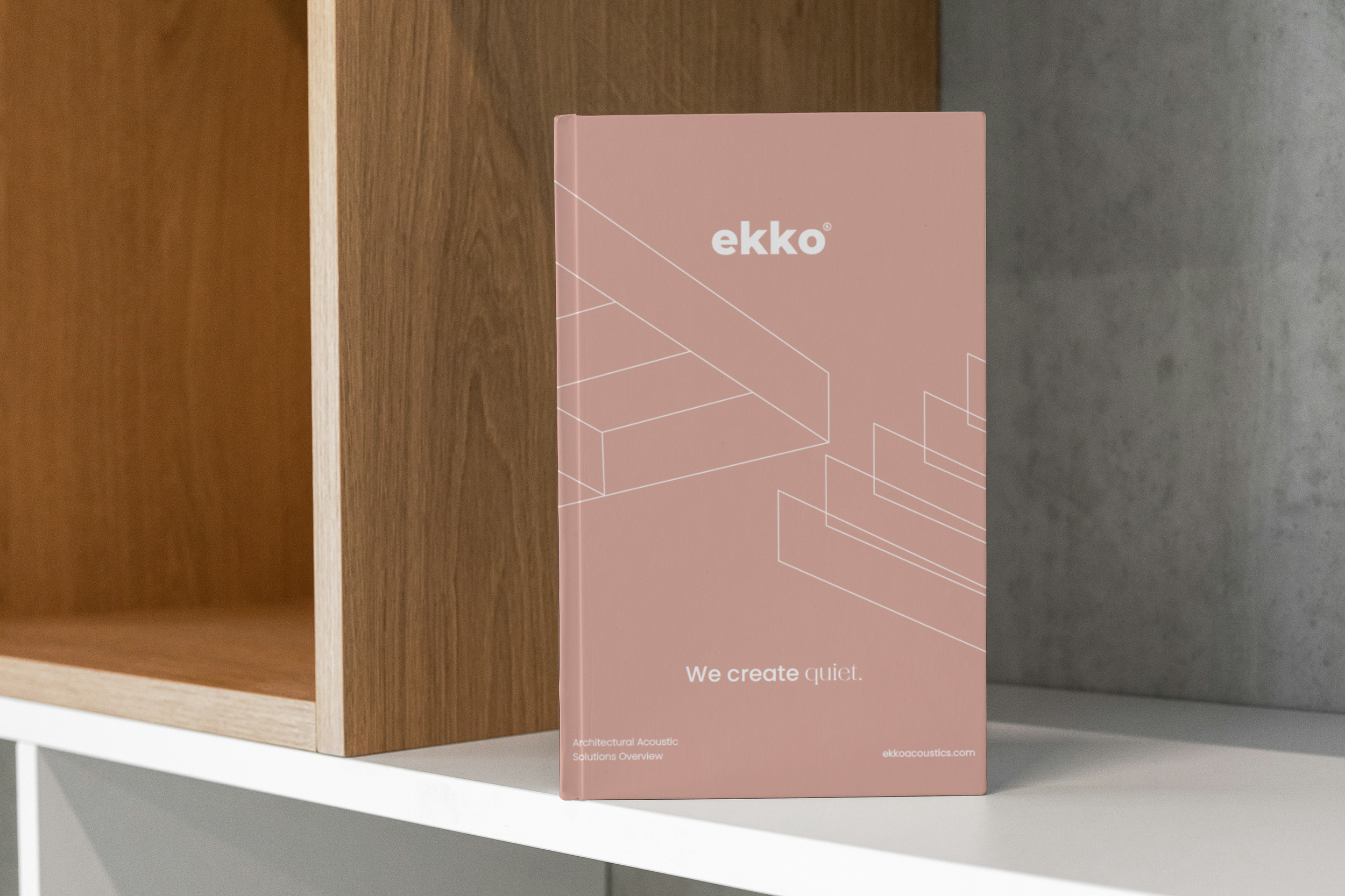 Pink brochure or booklet with the text 'ekko' and 'We create quiet.' on the cover, placed on a white shelf next to a wooden and concrete wall.