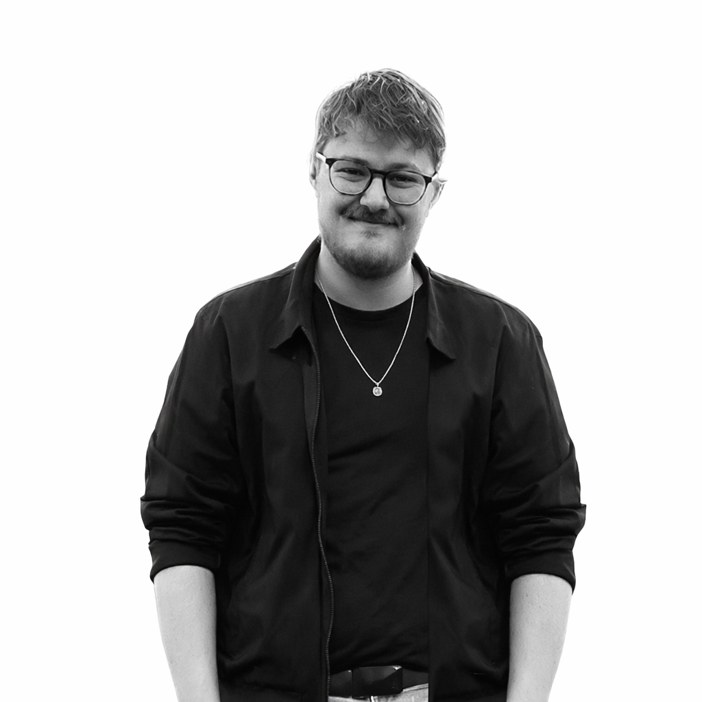 A young man with glasses, a beard, and a mustache wearing a black jacket, black t-shirt, necklace, and belt, standing against a plain white background.