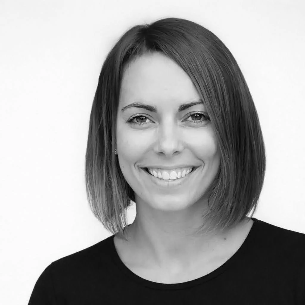 Black and white portrait of a young woman with straight, shoulder-length hair, smiling, wearing a dark top, and standing against a plain background.