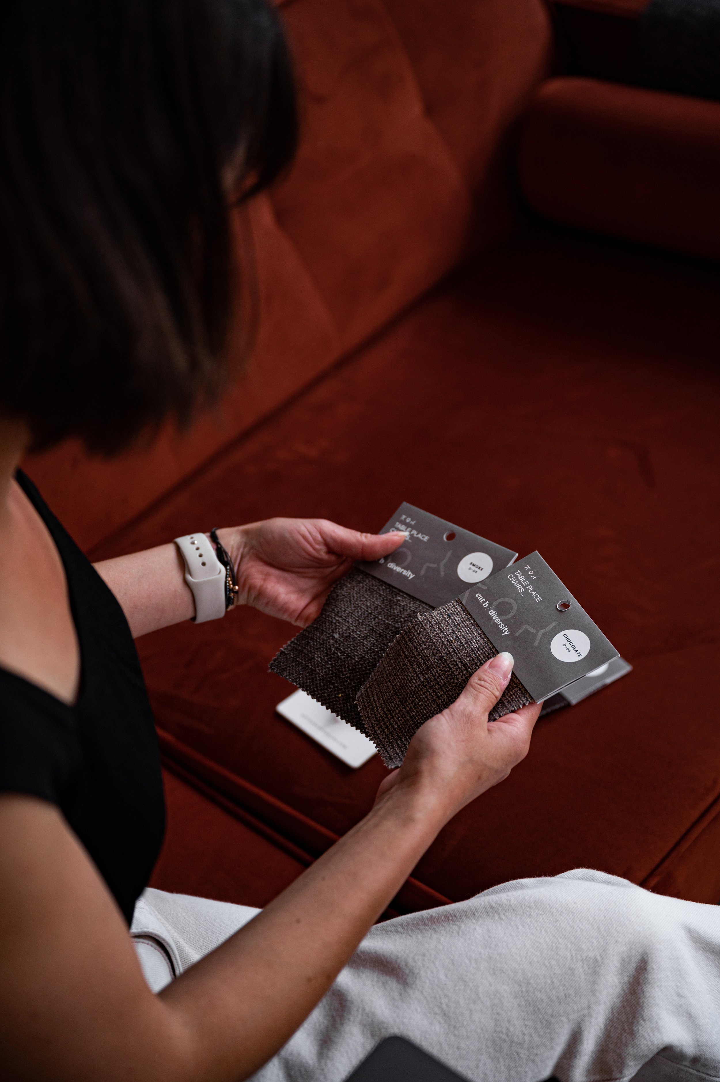 A woman sitting on a red sofa holding fabric swatches and color sample cards in her hands.
