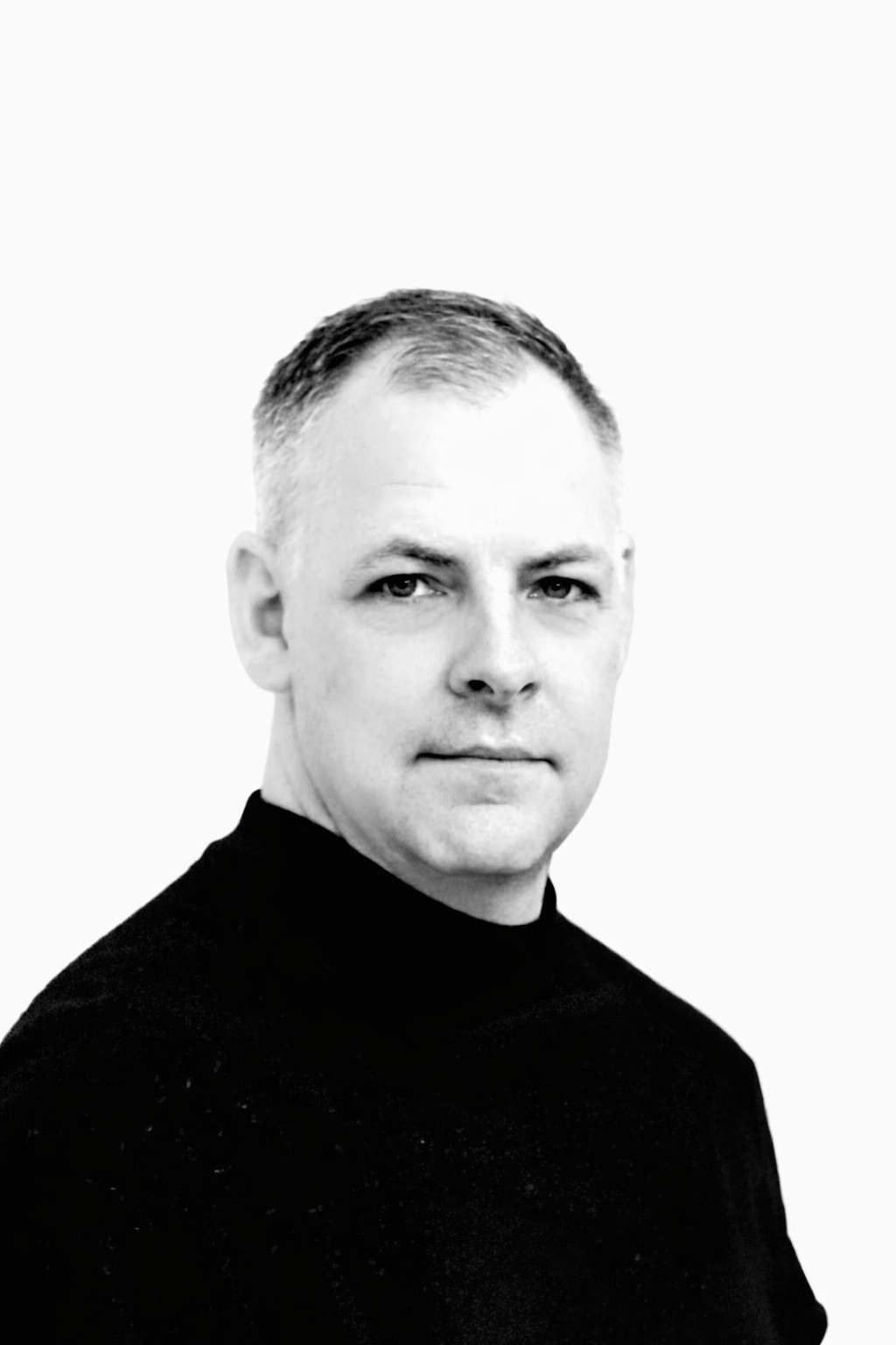 Black and white portrait of a man with short hair, wearing a black turtleneck, looking at the camera against a plain white background.