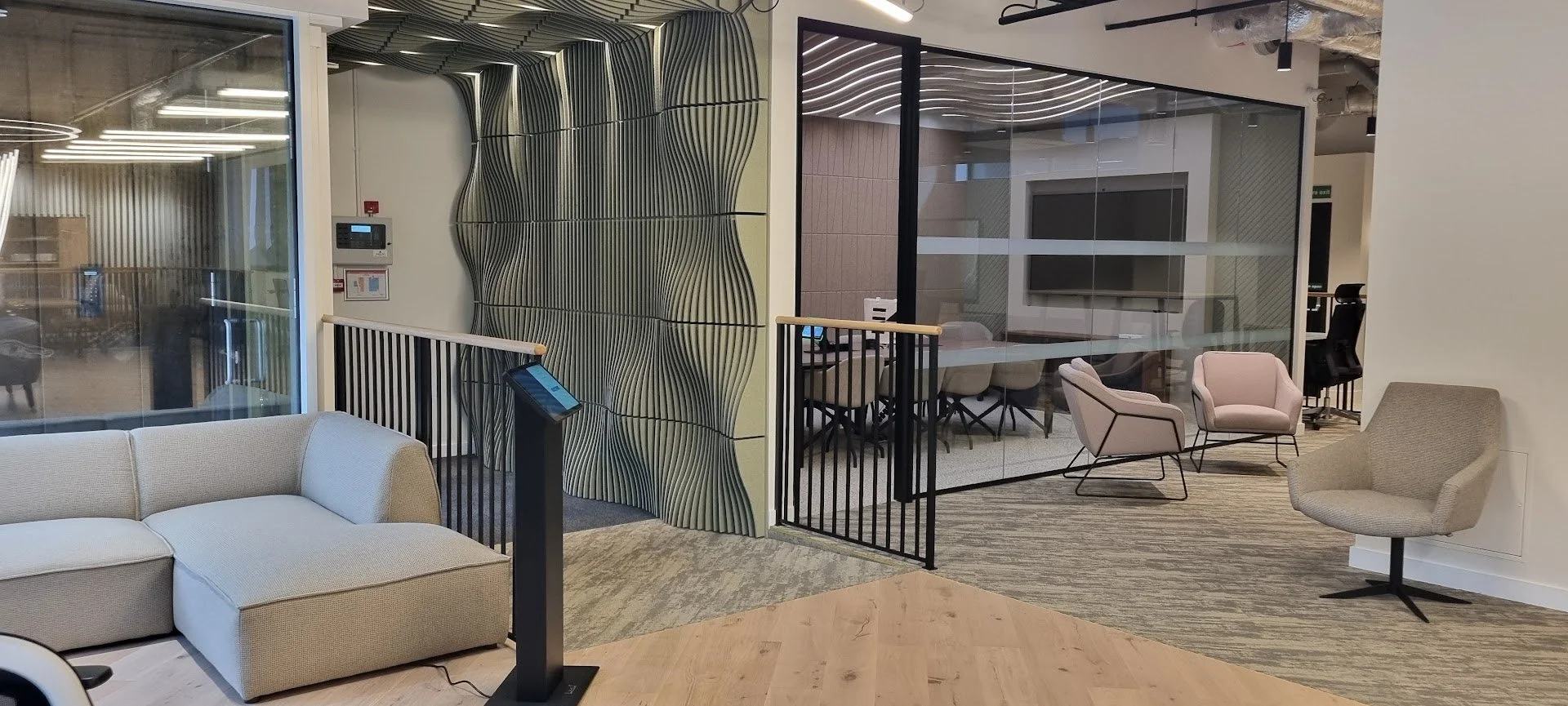 Modern showroom entrance area with a beige sectional sofa, pink and gray armchairs, and a glass enclosed meeting room with a large television.