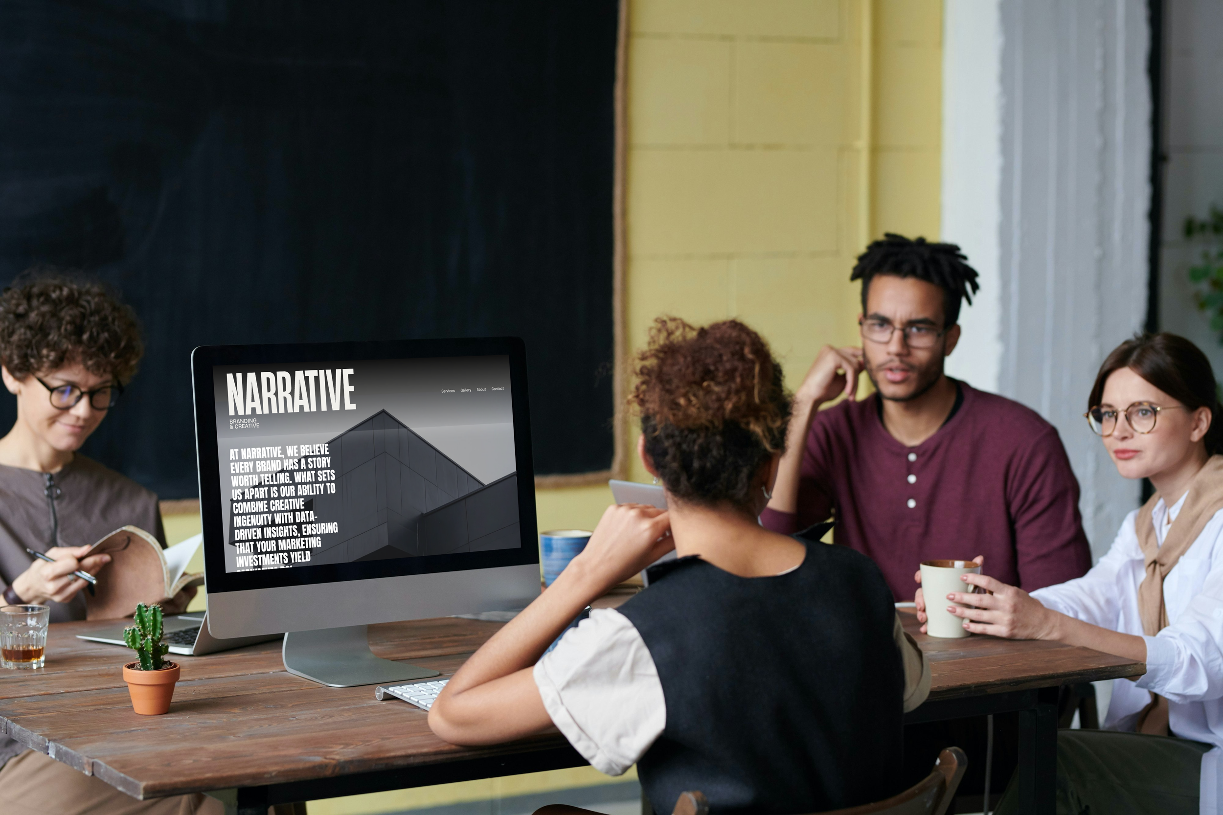 A group of five diverse people sitting around a wooden table having a discussion in a modern office with a yellow wall. One person is working on a large computer monitor, and others are drinking coffee or note-taking.
