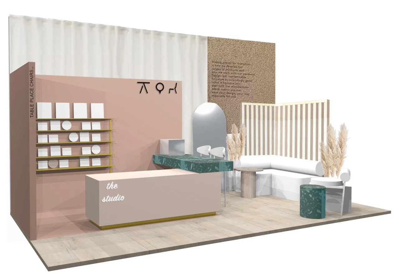A modern exhibition stand display with a pink wall, white sofa, gold shelving, vanity mirror, and decorative dried grasses, labeled as "the studio" with a welcome desk.