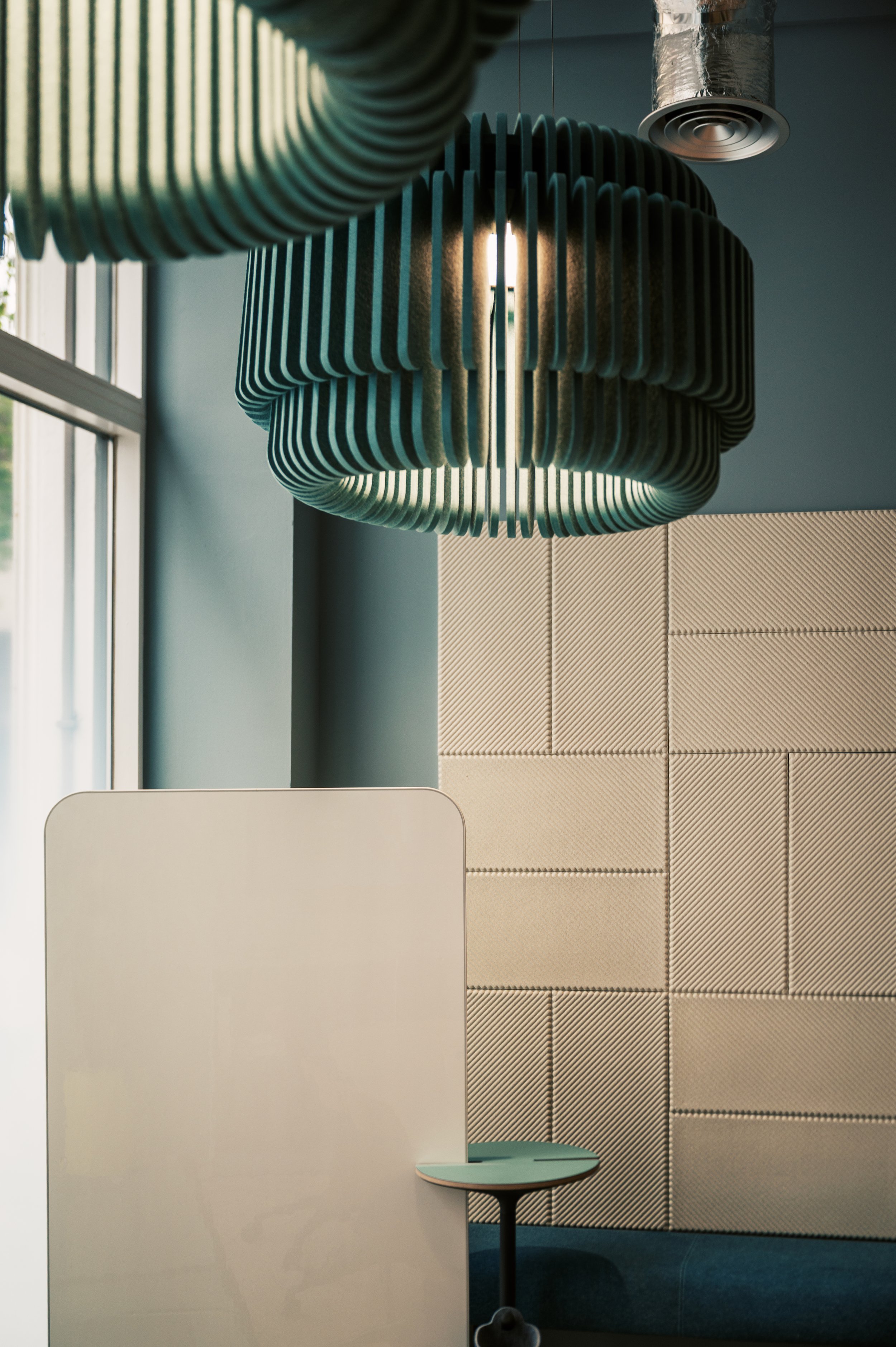 Modern interior space featuring a large, suspended, ribbed, circular light fixture, a beige tiled wall, a white partition or wall, a small round side table with a teal or light blue top, and a window with vertical glass panes.