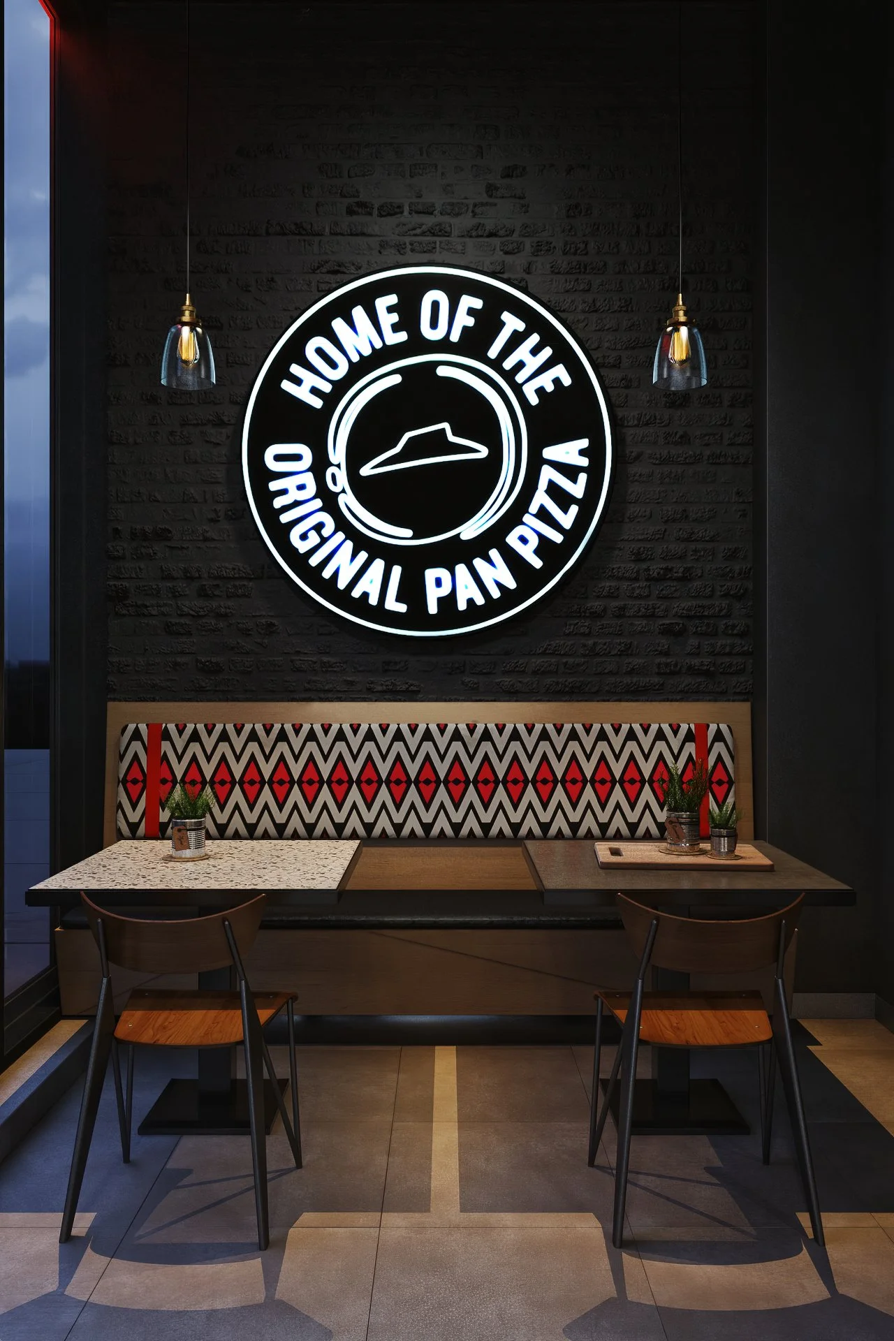 Interior of a pizza restaurant with a neon sign that says 'Home of the Original Pan Pizza' and a stylized pizza slice logo. There are two wooden chairs in front of a black marble table with potted plants, and a decorative booth with a zigzag patterne