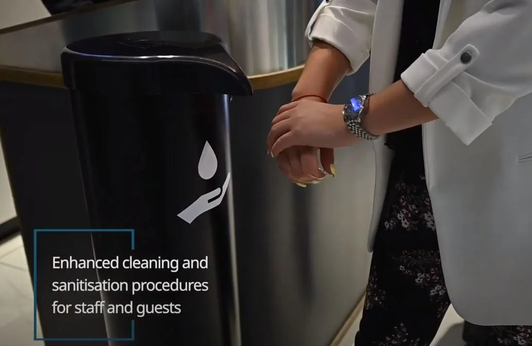 Person washing hands at a hand sanitizer station with a sign for enhanced cleaning and sanitization procedures.