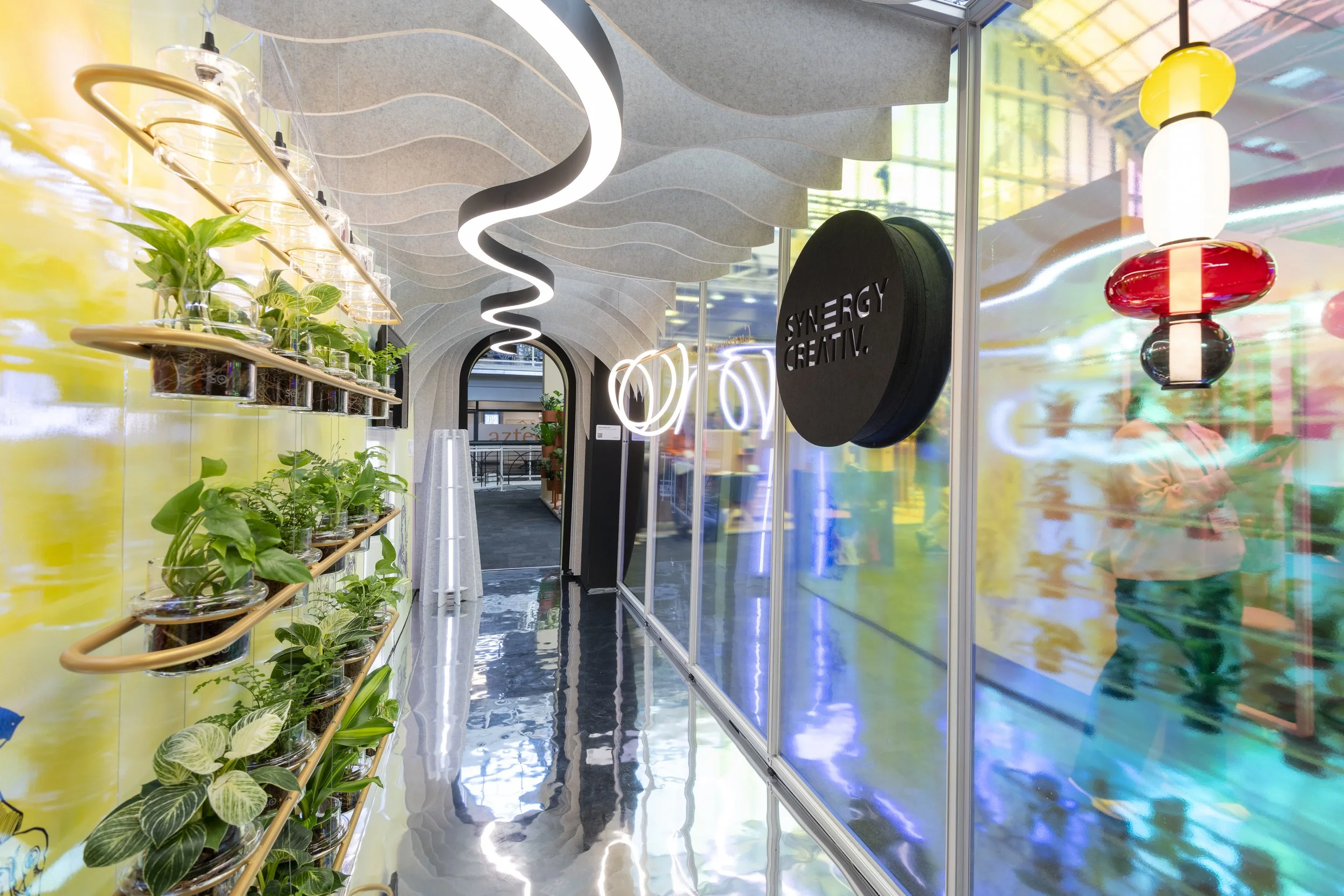 Exhibition stand with curved ceiling design and hanging plants on the left; colorful lighting fixtures on the right, and a sign that reads "SYNERGY CREATIV." in the middle.
