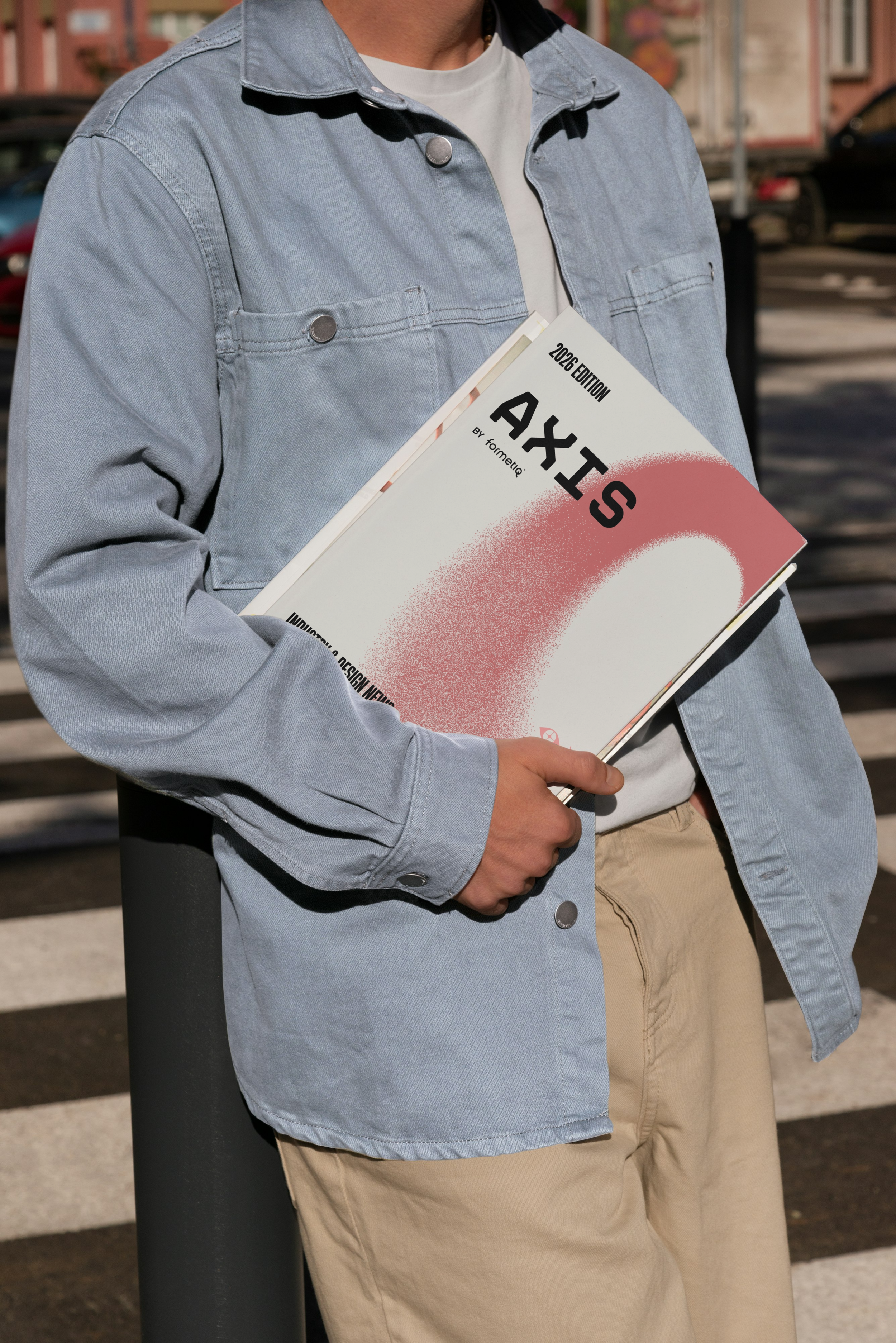 Person holding a large branded art magazine titled 'AXIS' with a pink gradient design, wearing a light blue denim jacket, beige pants, and a white t-shirt, standing outdoors near a crosswalk.