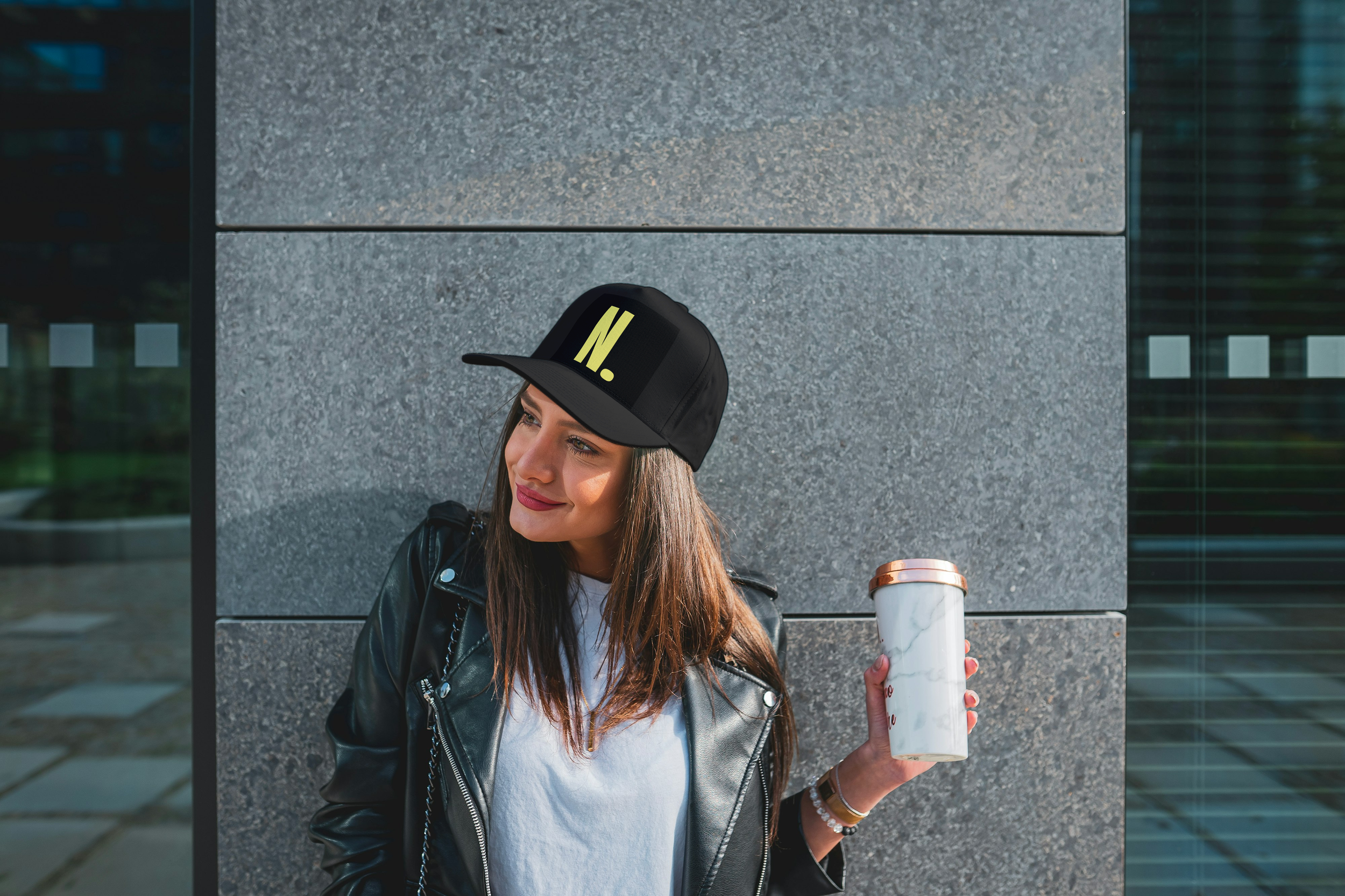A young woman with long brown hair, wearing a black leather jacket, white shirt, and a black cap with the letter 'N' on it, holding a marble-patterned travel mug and standing in front of a gray stone wall outside.