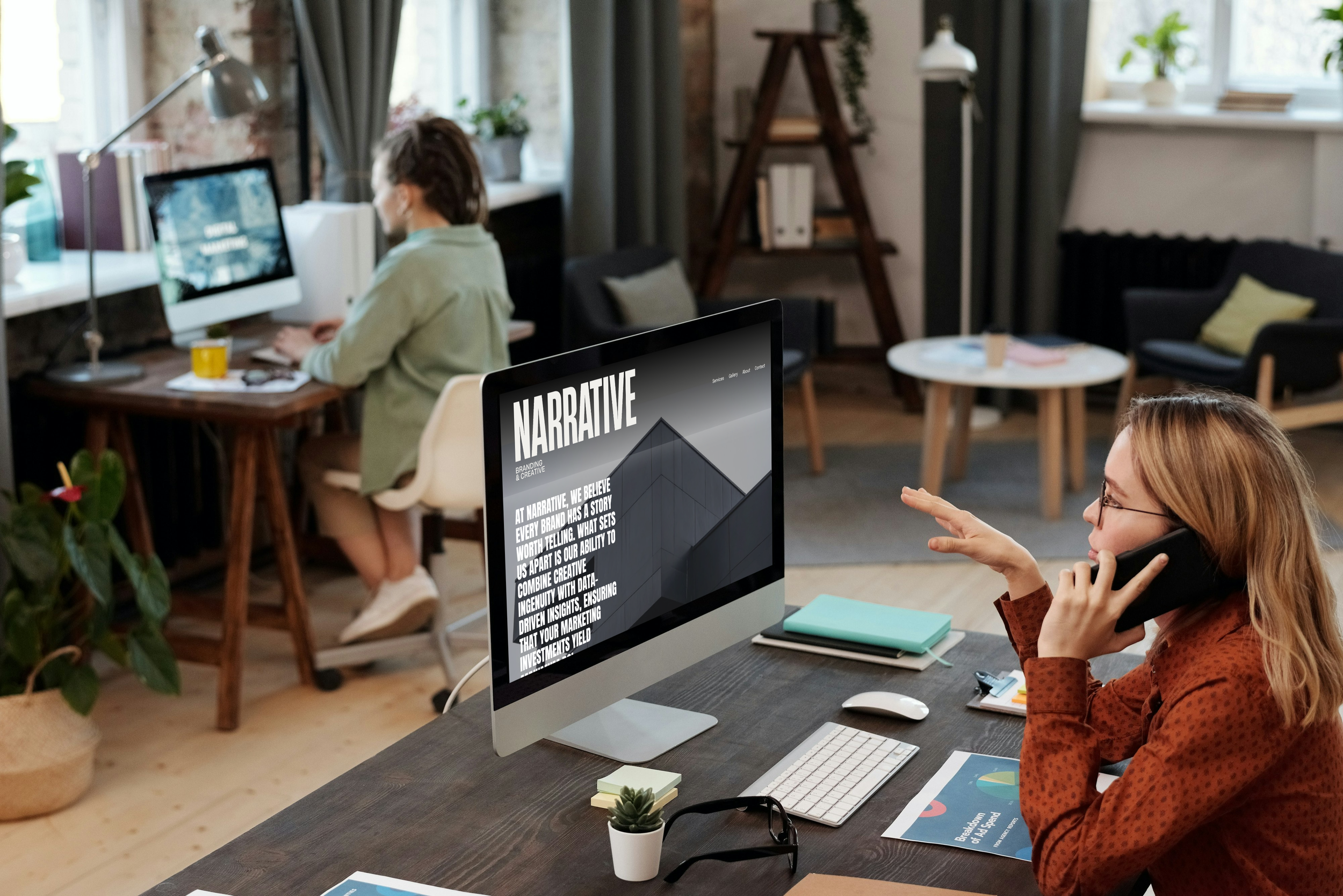 A woman with glasses and red hair talking on the phone at her desk in a modern office, with a computer displaying a presentation about 'Narrative'. In the background, a man is working on a computer at a separate desk.