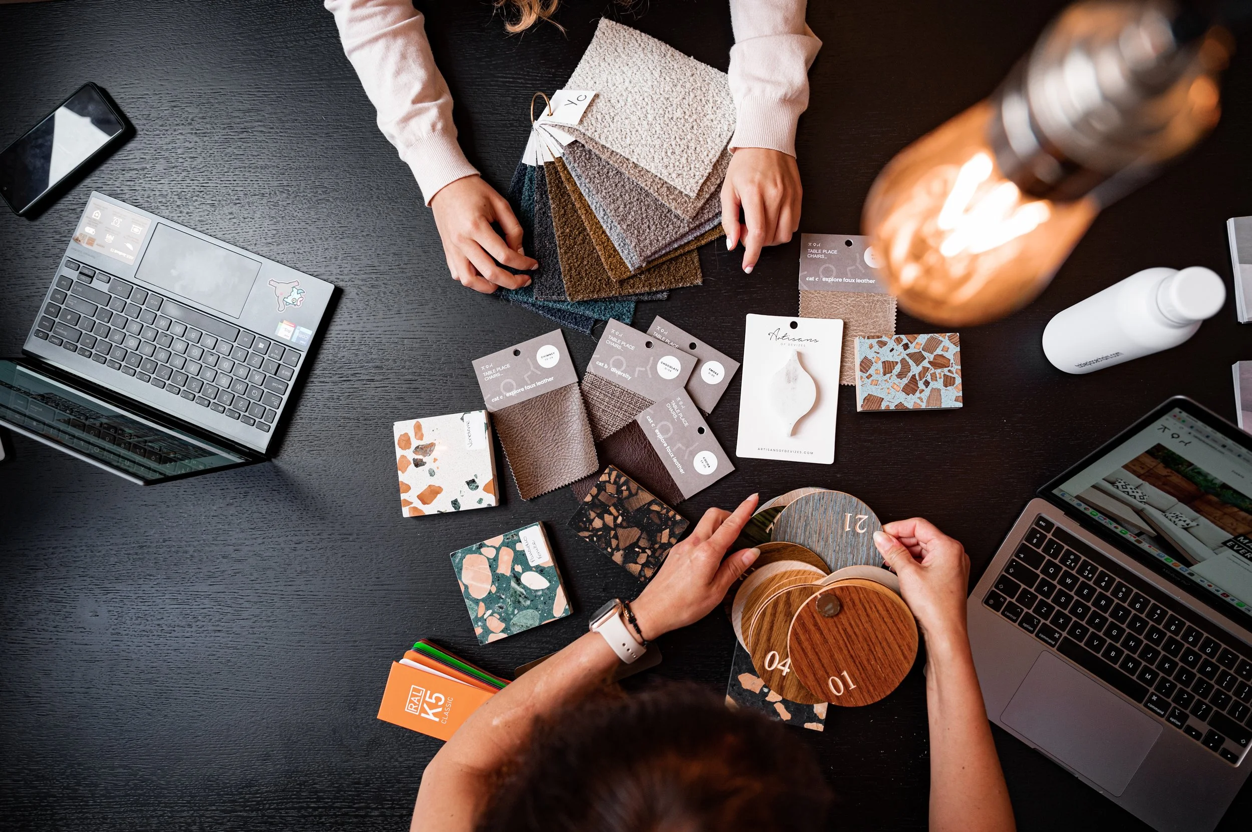 Two people organizing wood and tile samples on a black table, with a laptop, smartphone, color swatches, and fabric samples around.