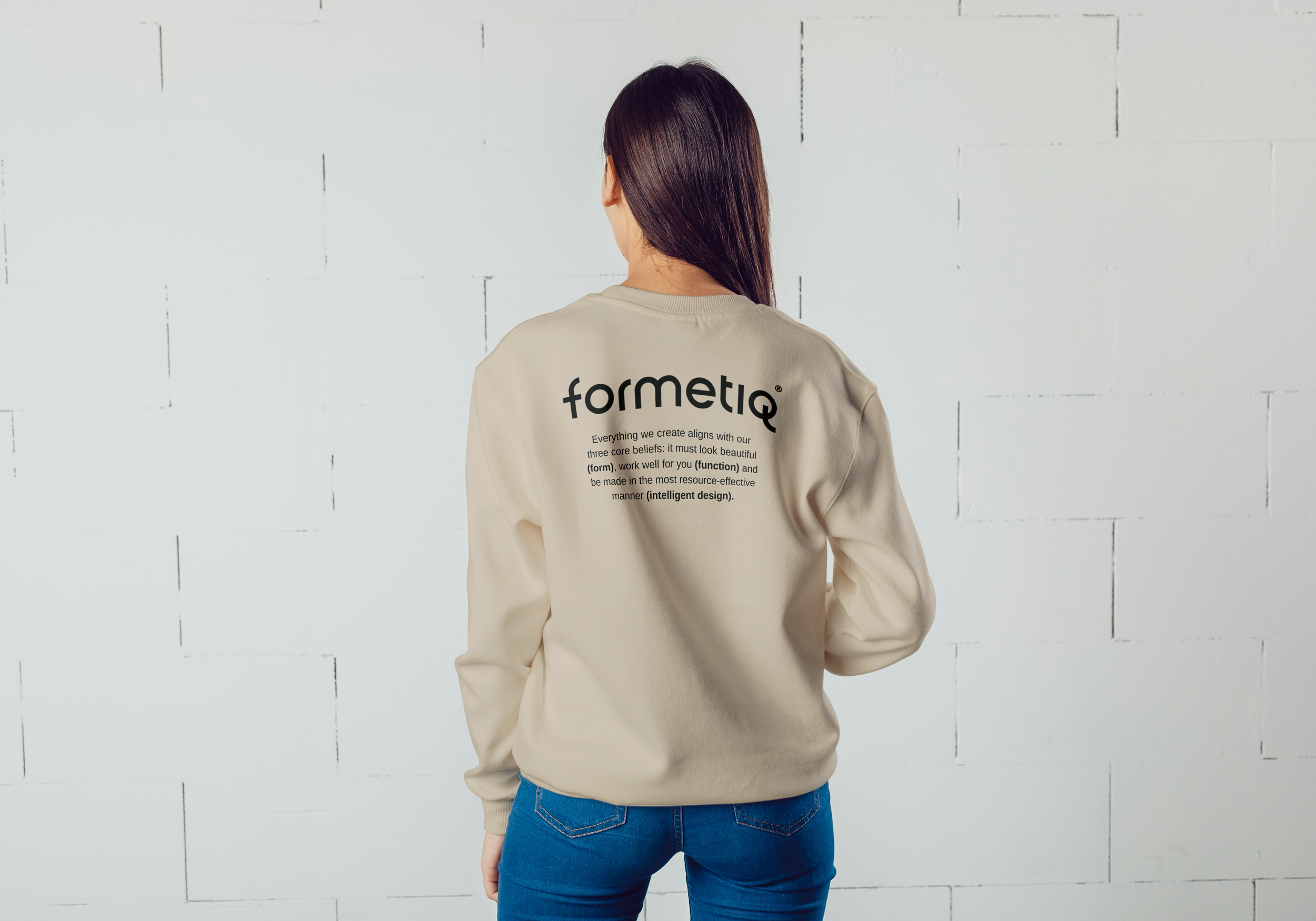 Back of a woman with long dark hair wearing a branded beige sweatshirt with black and grey text on the back, standing in front of a white brick wall.