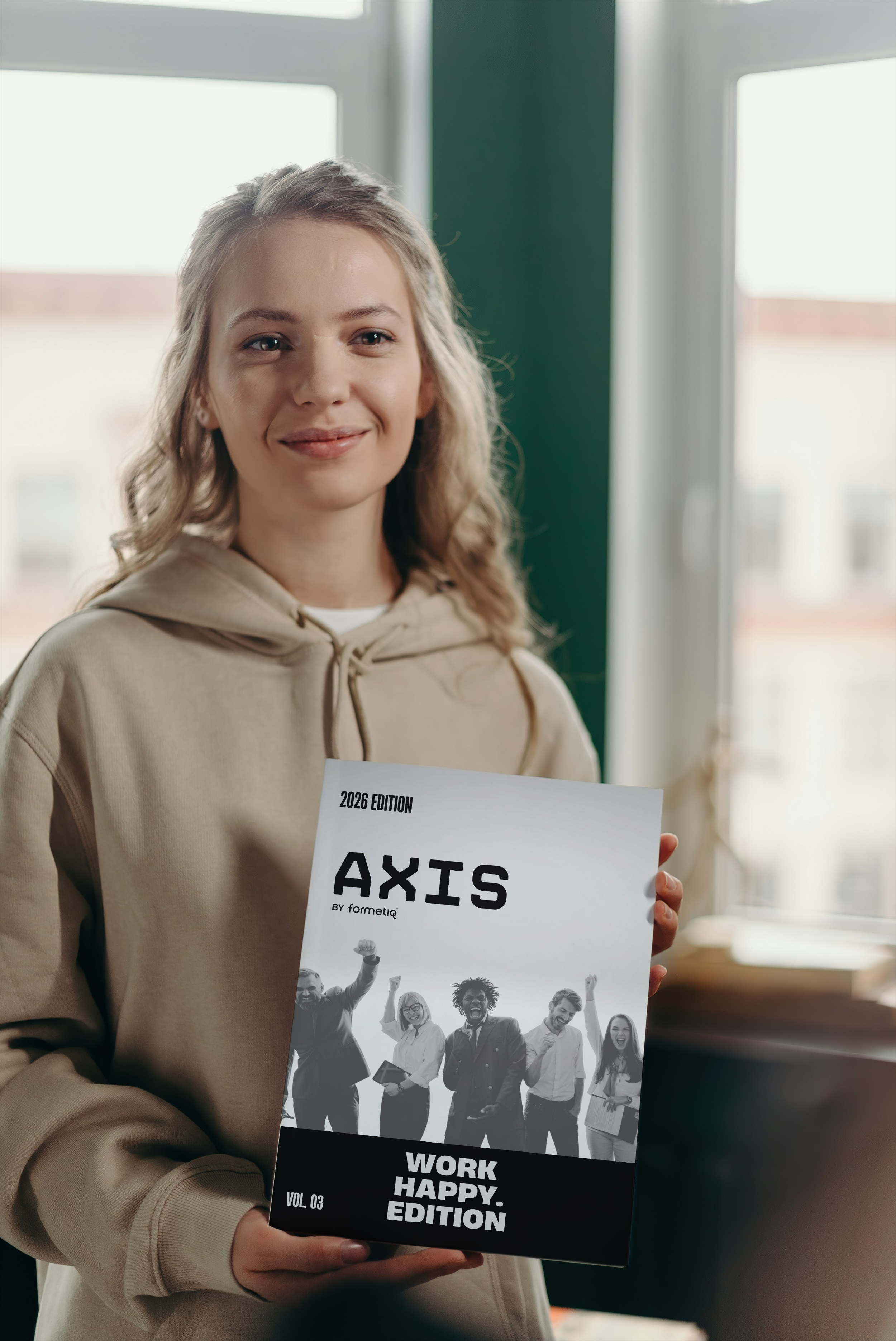 A young woman with curly blonde hair smiling and holding a magazine titled 'AXIS Work Happy Edition.' She is wearing a beige hoodie and standing near a window with a cityscape in the background.