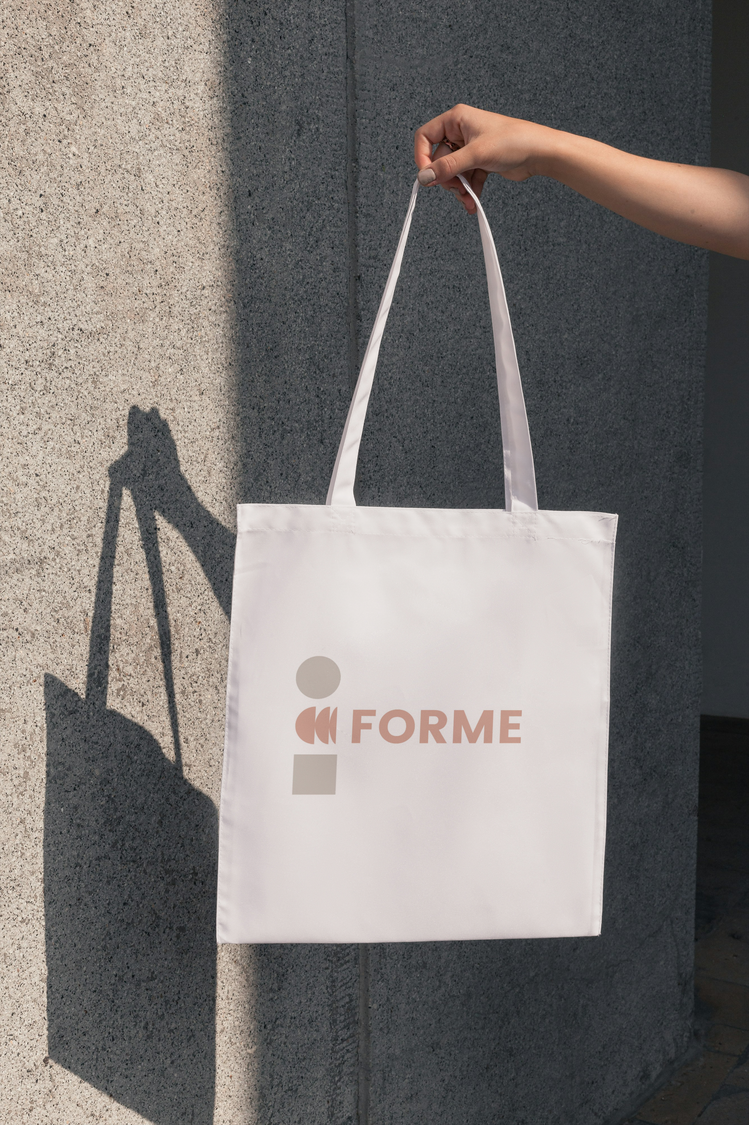 Close-up of a white tote bag with the logo and text 'i FORMÉ' held by a person's hand against a textured wall, with a shadow cast on the wall.