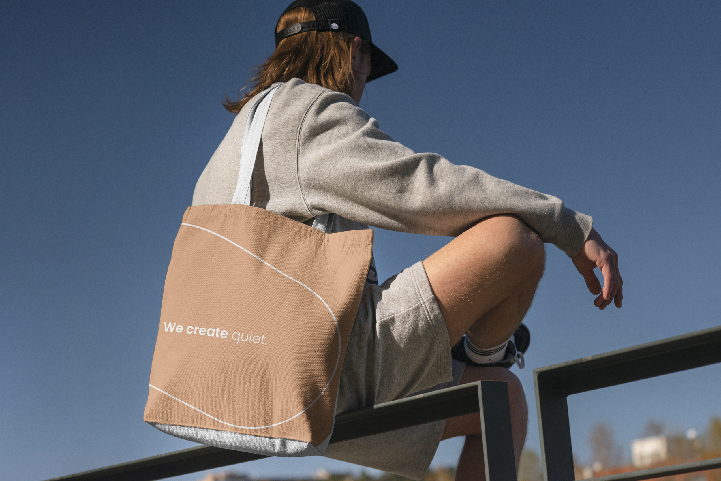 Person with long hair wearing a black cap, gray sweatshirt, and shorts, sitting on a railing with a tan tote bag that says "We create quiet." against a clear blue sky.