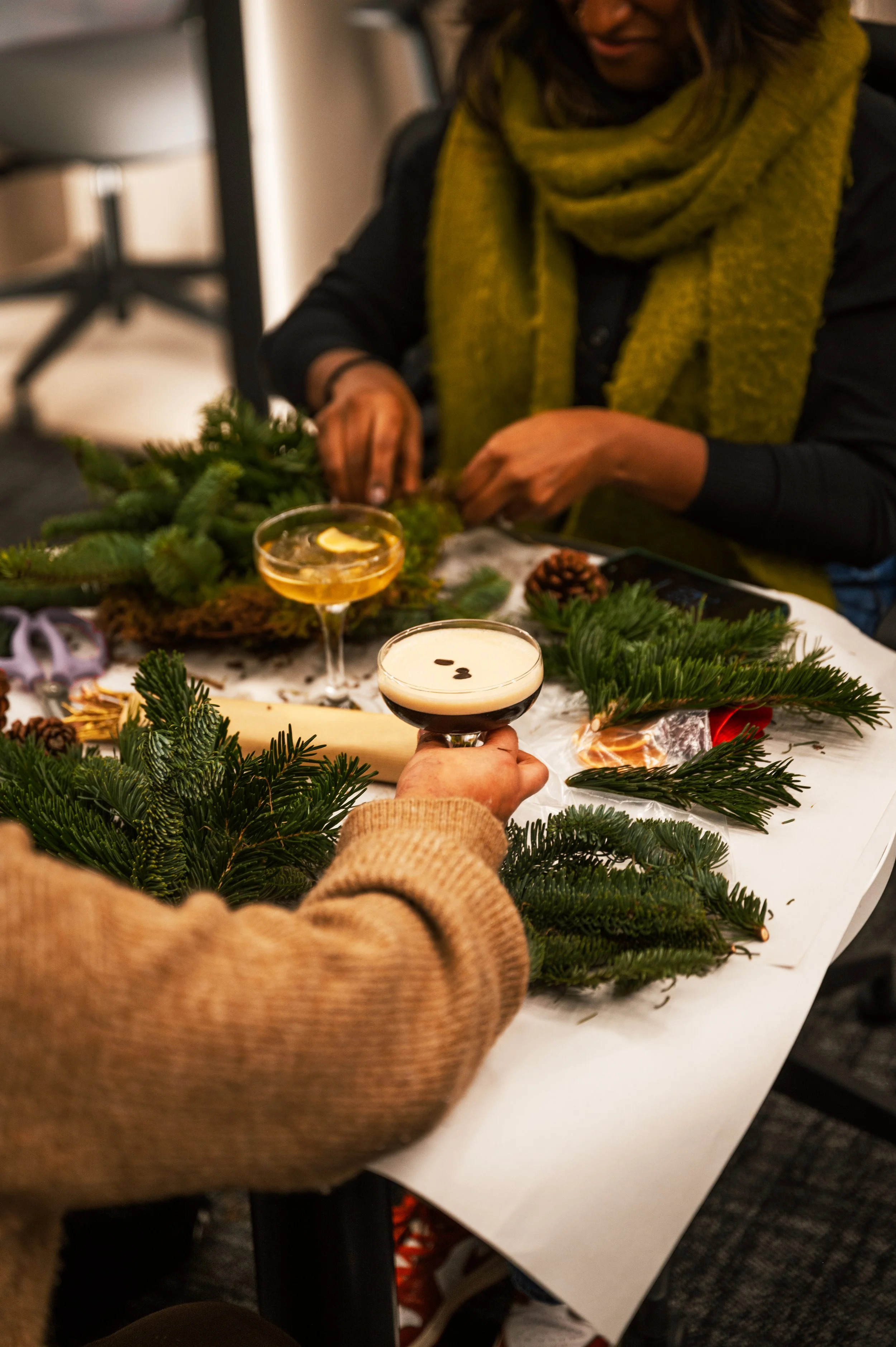 Two people are decorating a table with pine branches and pinecones, with drinks in hand during a festive gathering.
