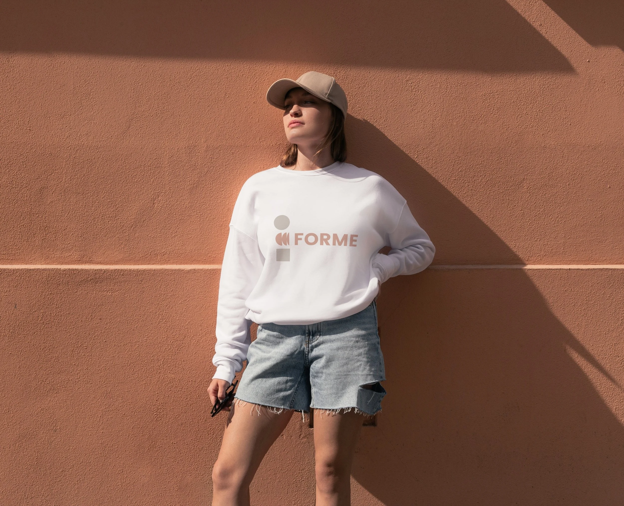 Young woman with a beige cap, white sweatshirt with the word 'FORME' and geometric design, gray denim shorts, standing against a pinkish wall with shadow.